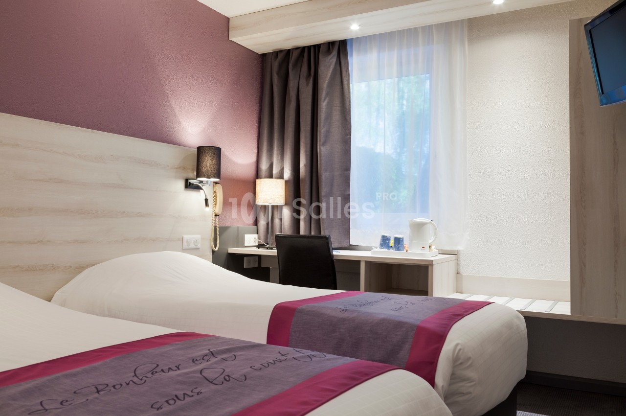 Chambre d'hôtel avec deux lits simples, bureau équipé, lampe, fenêtre avec rideaux et décoration sobre.