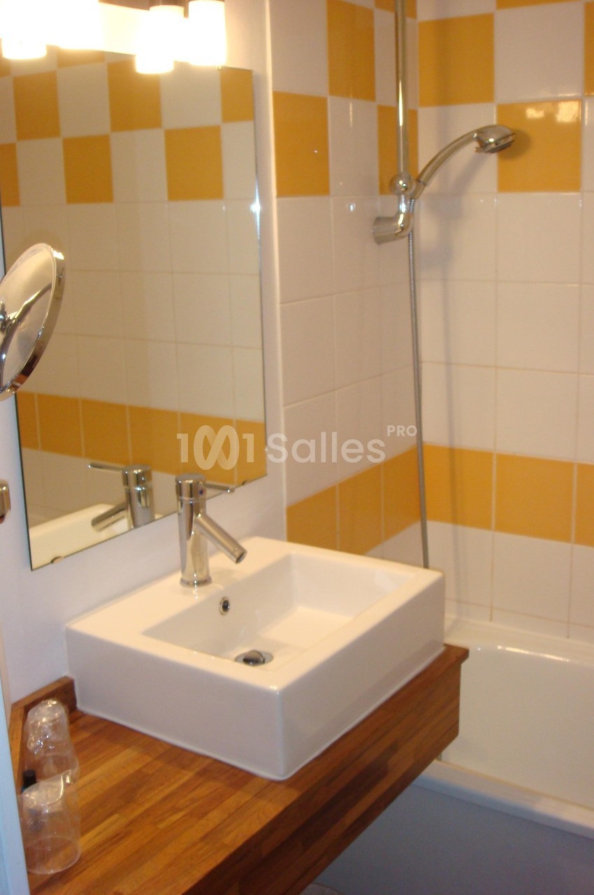 Lavabo blanc sur un plan de travail en bois, avec miroir, douche et carrelage blanc et jaune en arrière-plan.