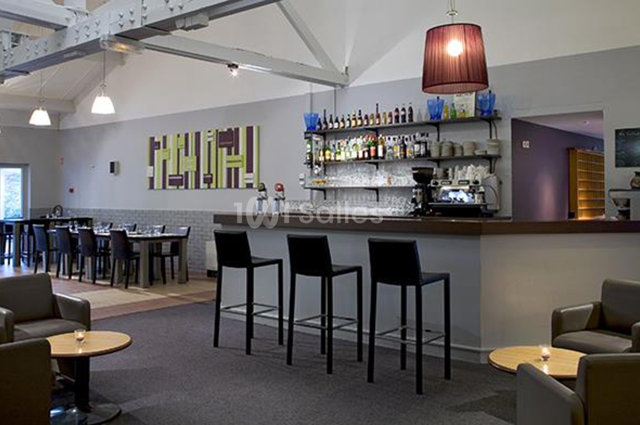 Bar moderne avec comptoir en bois, tabourets hauts, étagères de bouteilles, et coin salon avec fauteuils et tables basses.