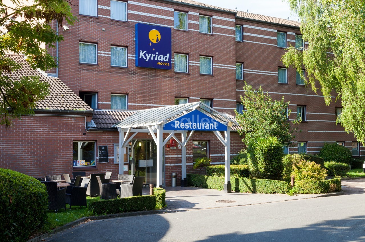 Façade d'un hôtel en briques rouges avec un panneau ’Kyriad Hotel’ et une entrée de restaurant entourée de verdure.