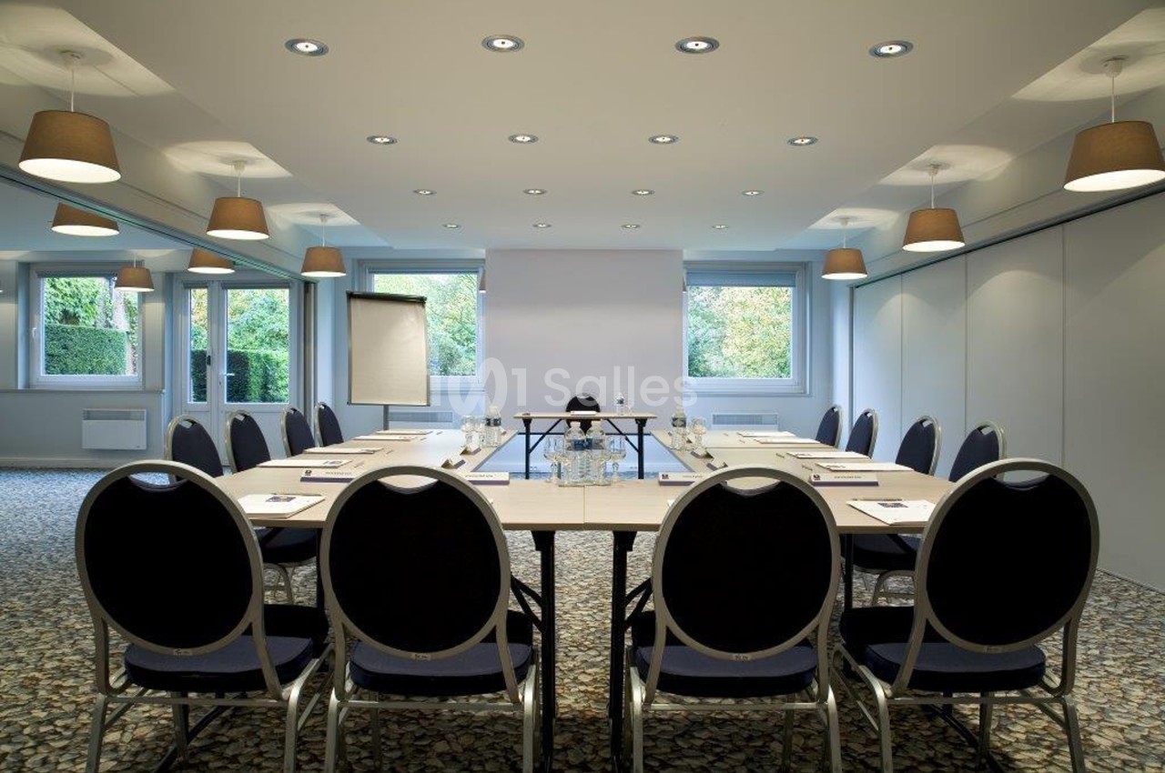 Salle de réunion lumineuse avec une grande table ovale, chaises noires, écran de projection et fenêtres donnant sur un…