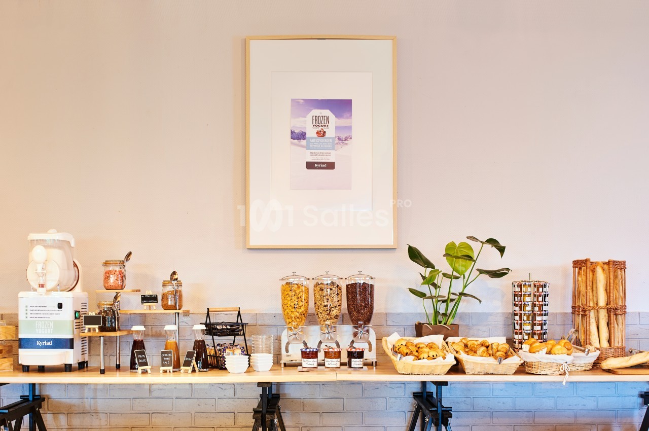 Buffet de petit-déjeuner avec céréales, pains, confitures, machine à jus et décorations murales simples.