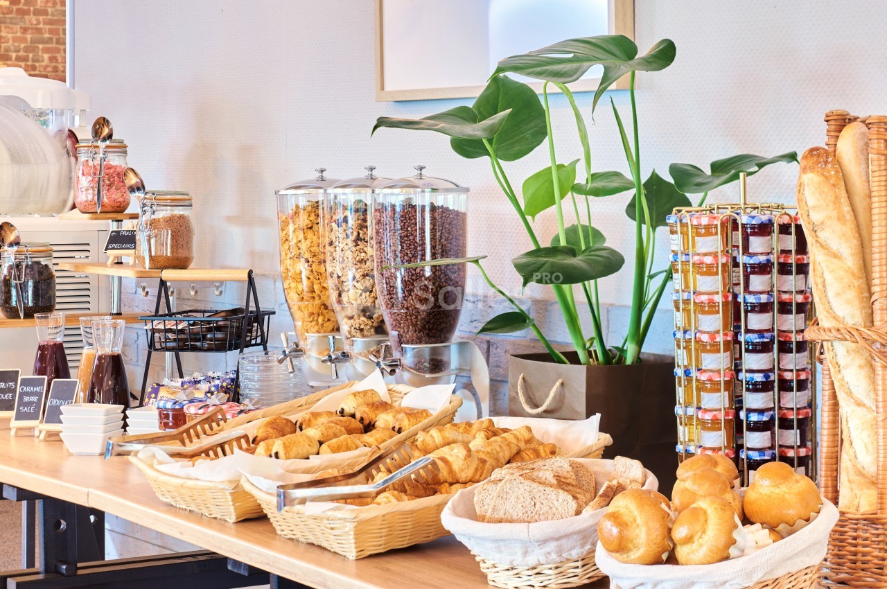 Buffet de petit-déjeuner avec viennoiseries, pains, confitures, céréales et machine à café dans un décor lumineux.