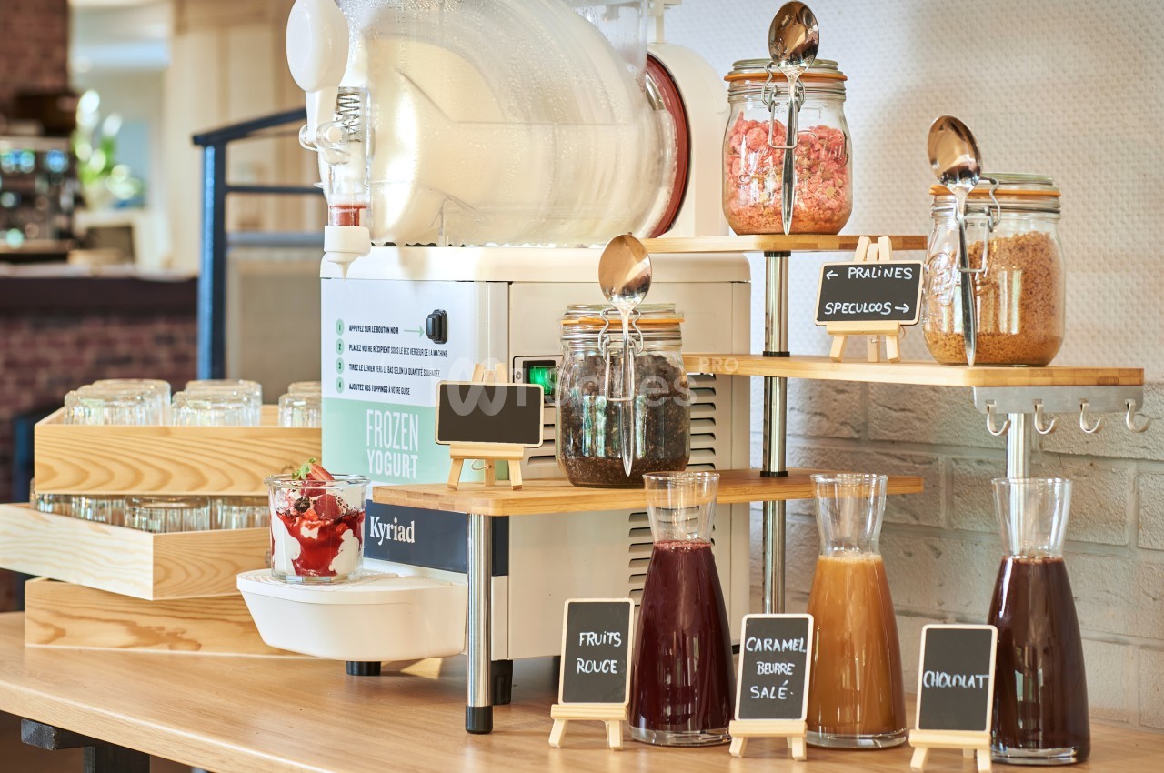 Machine à yaourt glacé avec garnitures variées en pots et carafes, disposées sur un comptoir en bois.