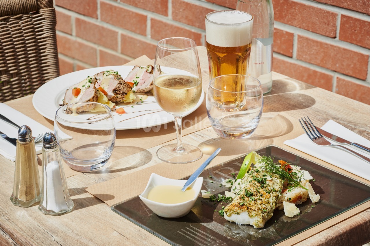 Repas servi en terrasse avec poisson, viande, accompagnements, verre de vin blanc et bière sur une table en bois.