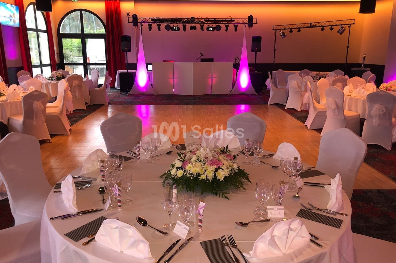 Salle de réception décorée avec des tables rondes dressées, nappes blanches et éclairage coloré en arrière-plan.