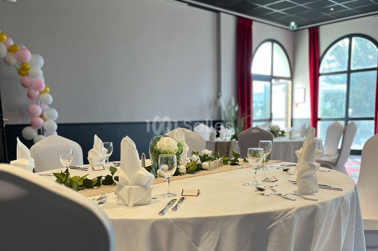 Table ronde dressée pour un événement, avec nappes blanches, serviettes pliées, verres et décoration florale.