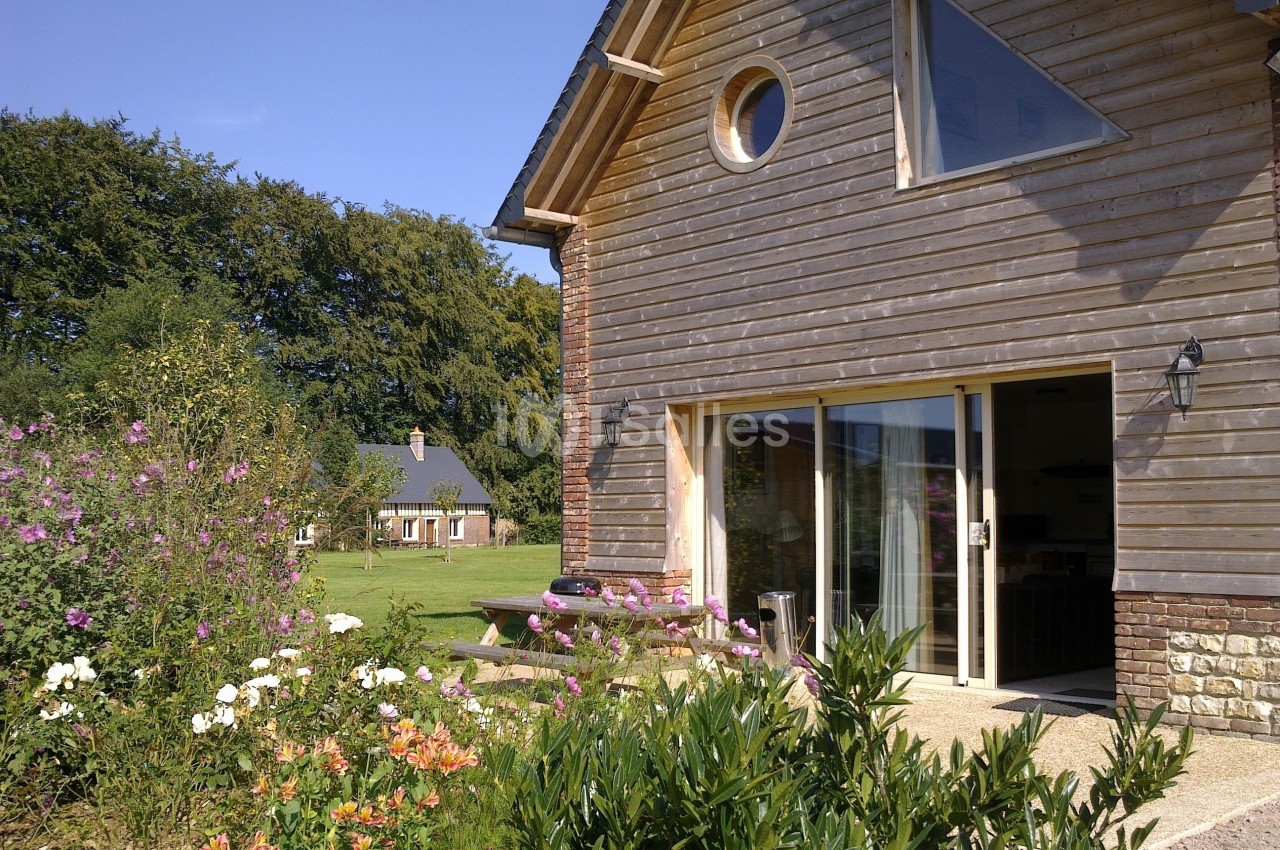 Maison en bois avec de grandes baies vitrées, entourée d'un jardin fleuri et d'arbres en arrière-plan.
