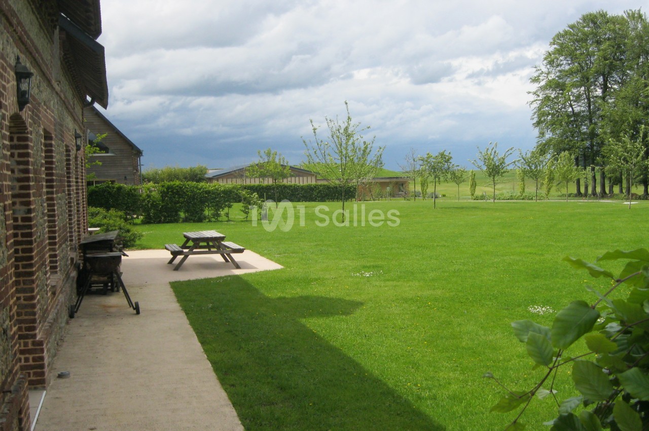 Jardin verdoyant avec pelouse, arbres jeunes, tables de pique-nique et bâtiments en briques sous un ciel nuageux.