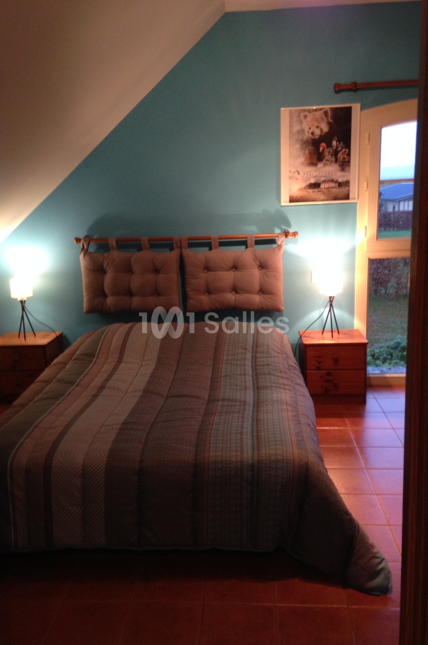 Chambre avec un lit double, mur bleu, deux tables de chevet en bois avec lampes et une fenêtre donnant sur un jardin.