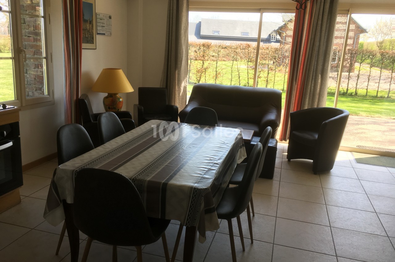 Salle à manger lumineuse avec table rectangulaire, chaises noires, canapé, fauteuils et vue sur un jardin par une baie…