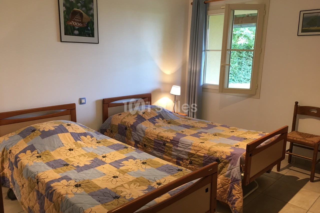 Deux lits simples avec couvre-lits fleuris dans une chambre lumineuse, une fenêtre et une chaise en bois.