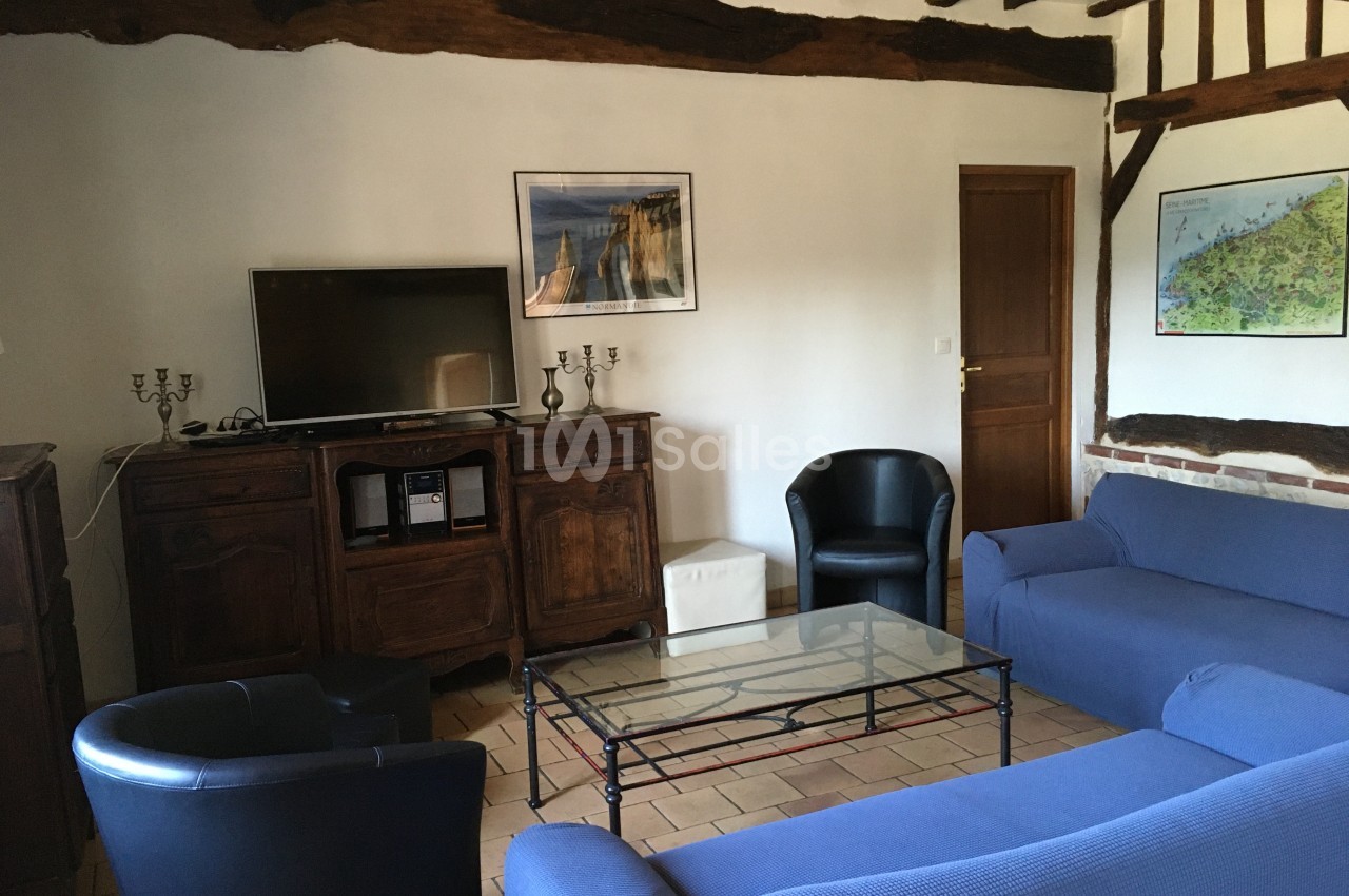 Salon avec poutres apparentes, canapé bleu, fauteuils noirs, table basse en verre et meuble TV en bois.