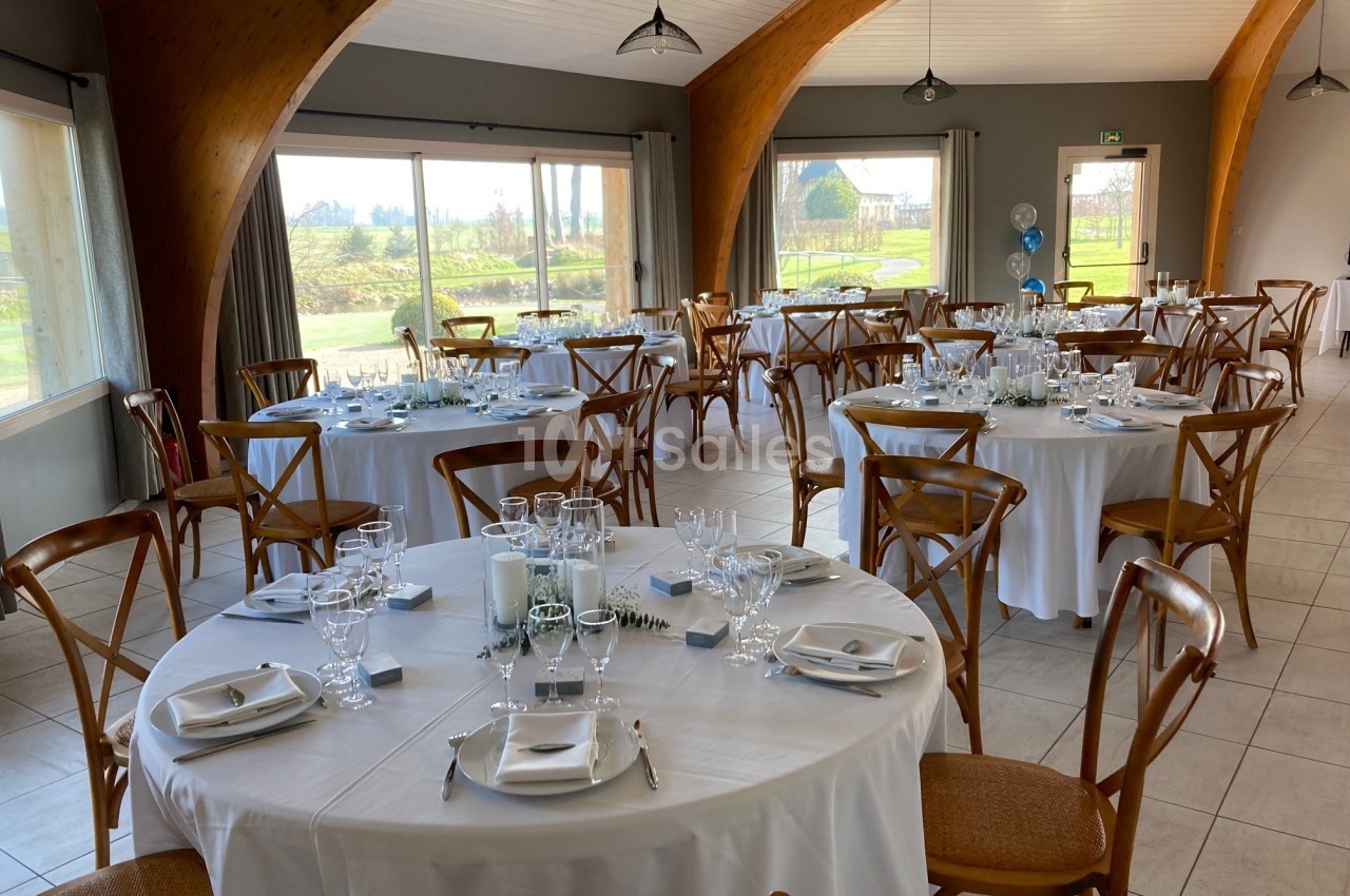 Salle de réception lumineuse avec tables rondes dressées, nappes blanches et chaises en bois, vue sur un paysage extérieur.
