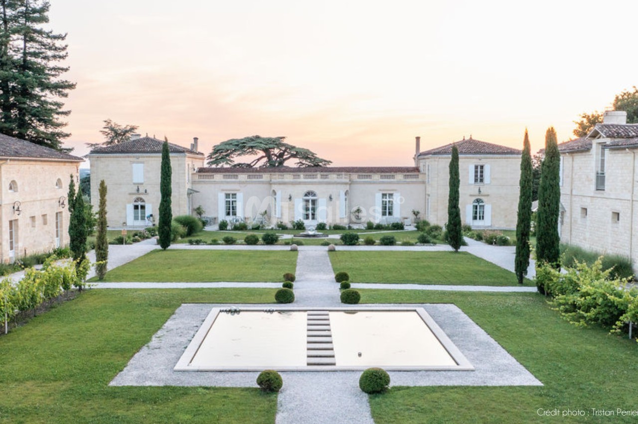 Vue d'un domaine avec jardin symétrique, bassin central, allées gravillonnées et bâtiments en pierre sous un ciel clair.