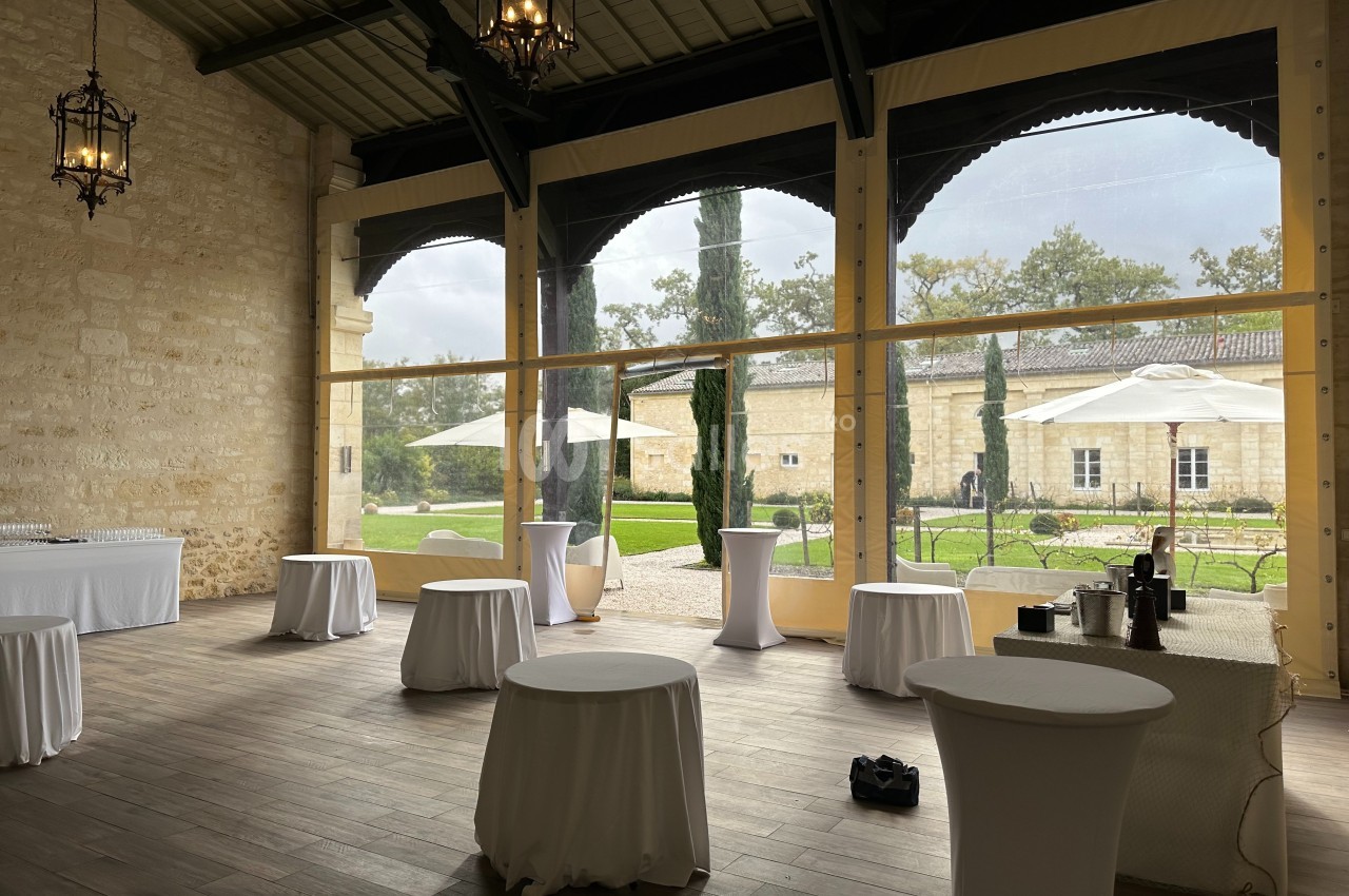 Salle lumineuse avec tables hautes drapées de blanc, grandes baies vitrées donnant sur un jardin avec parasols.