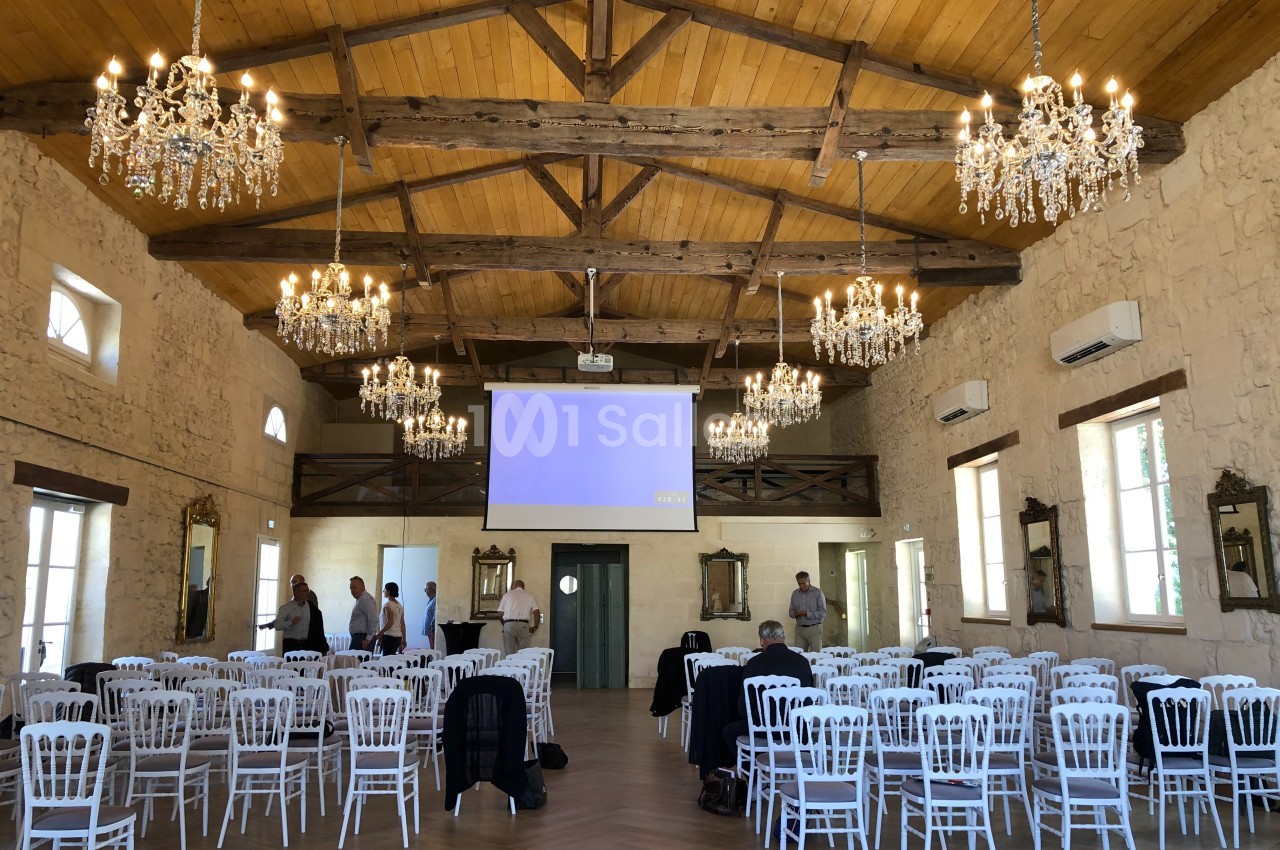 Salle de réception avec poutres apparentes, lustres, chaises blanches alignées et écran de projection allumé.