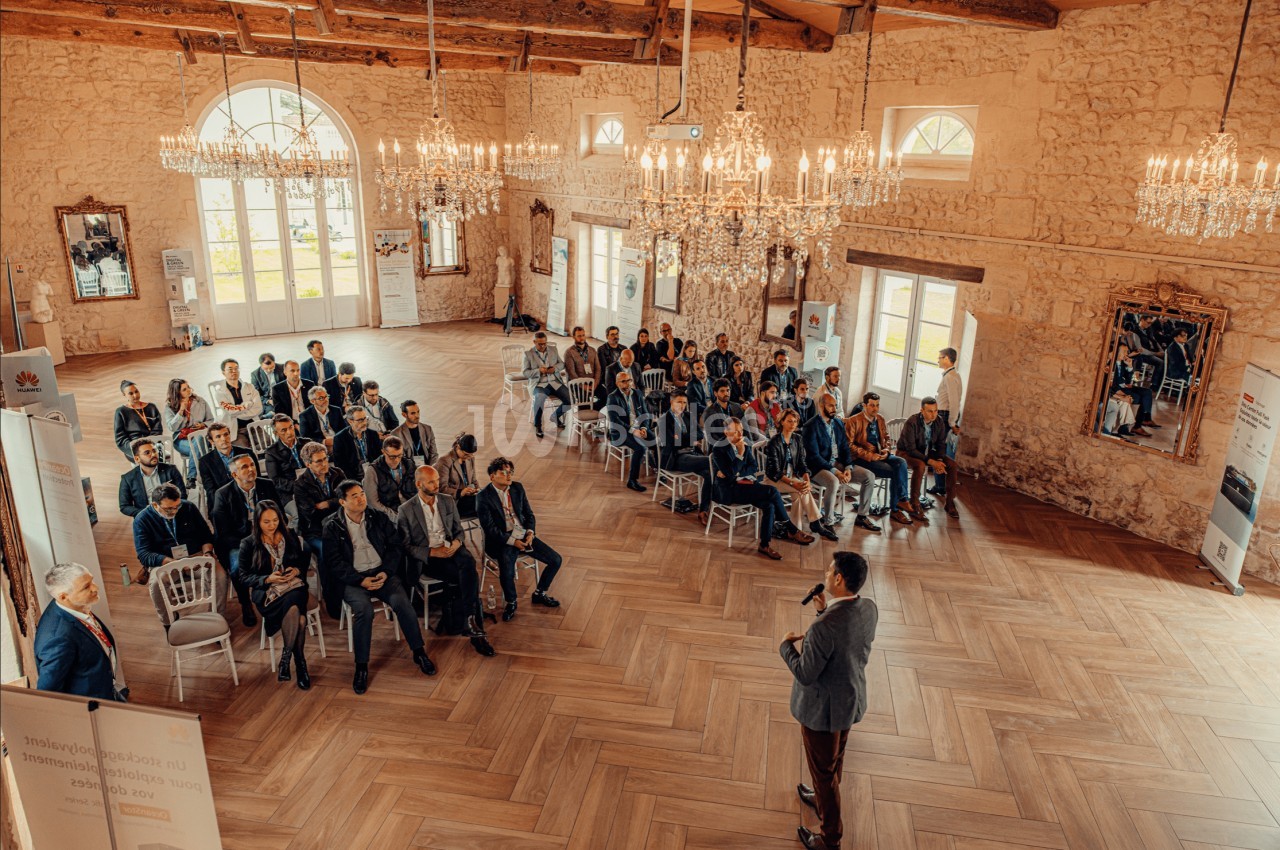 Un conférencier s'adresse à un public assis dans une salle lumineuse avec des murs en pierre et des lustres.