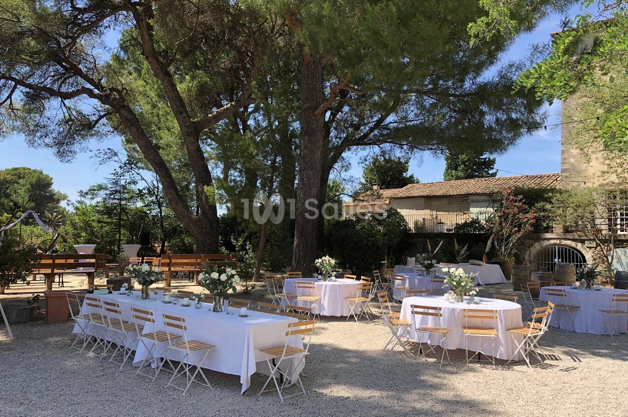 Tables dressées avec nappes blanches et chaises pliantes disposées en extérieur sous des arbres, dans une cour ensoleillée.