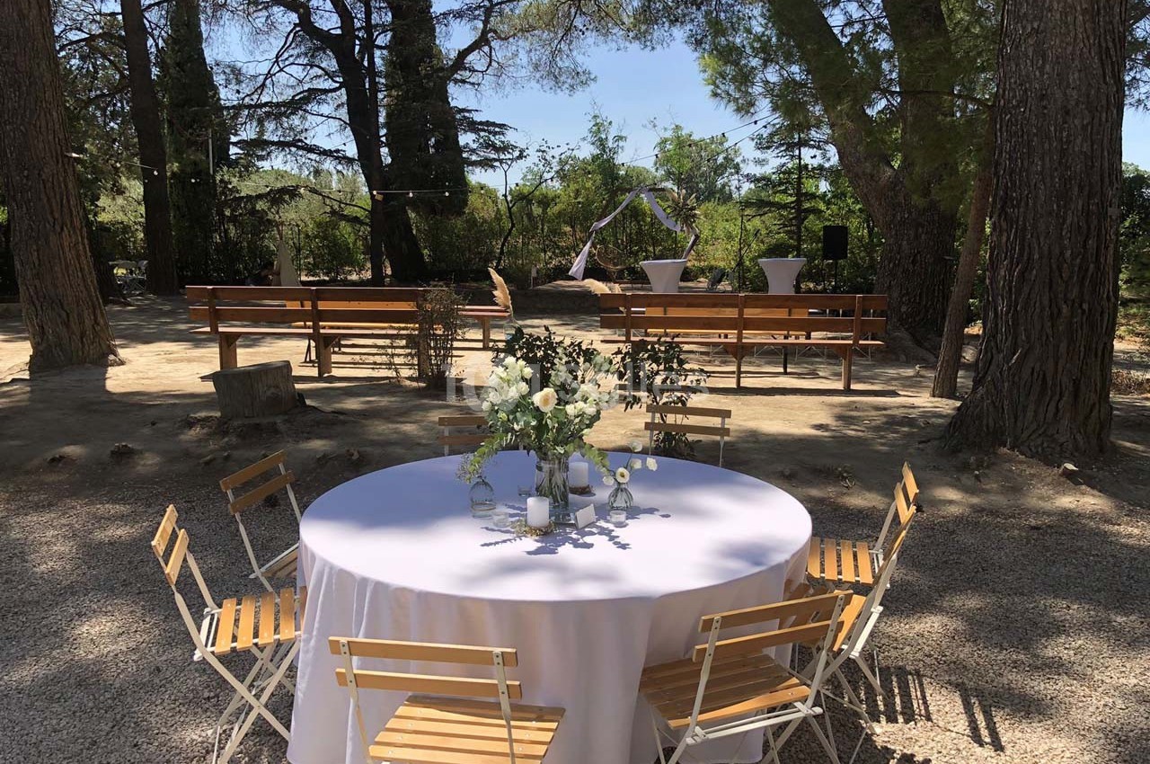 Table ronde décorée avec des fleurs et des chaises en bois, installée en extérieur sous des arbres, avec des bancs en…