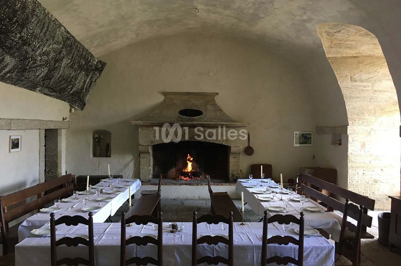 Salle à manger rustique avec tables dressées, cheminée en pierre allumée et murs en pierre apparente.