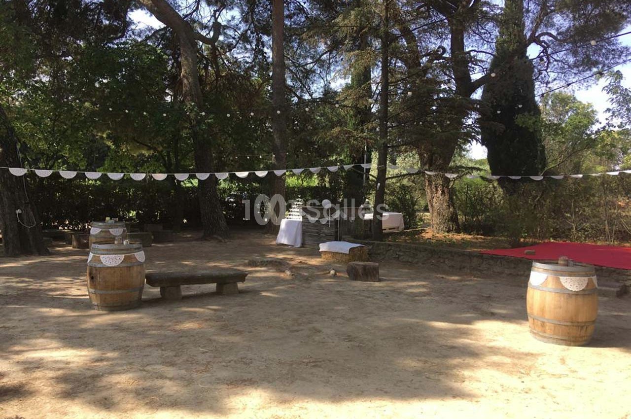 Espace extérieur ombragé avec guirlandes, tonneaux en bois, bancs rustiques et table blanche décorée.