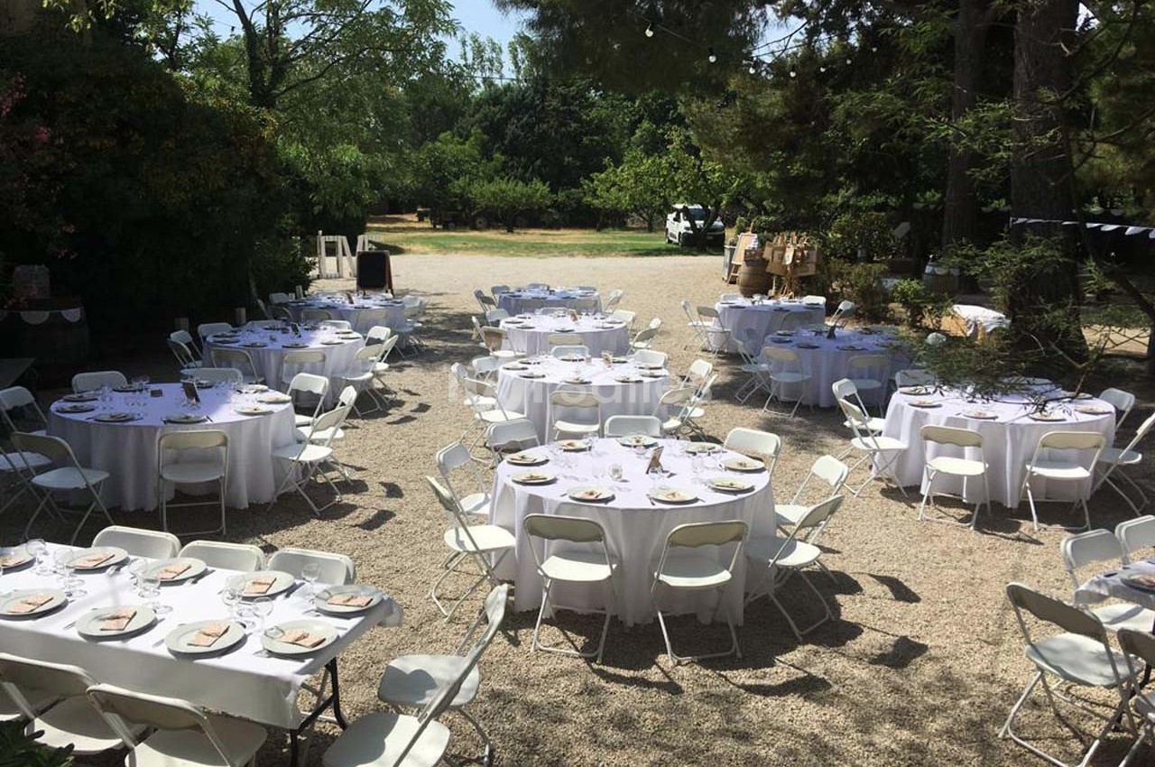 Tables rondes dressées avec nappes blanches et chaises pliantes dans un espace extérieur ombragé.