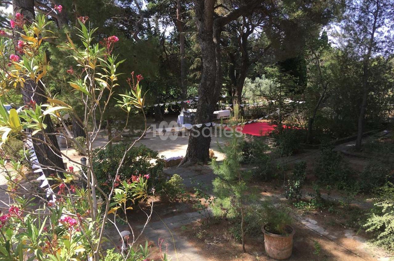 Jardin ombragé avec des arbres, des plantes fleuries, des guirlandes et des tables dressées en arrière-plan.