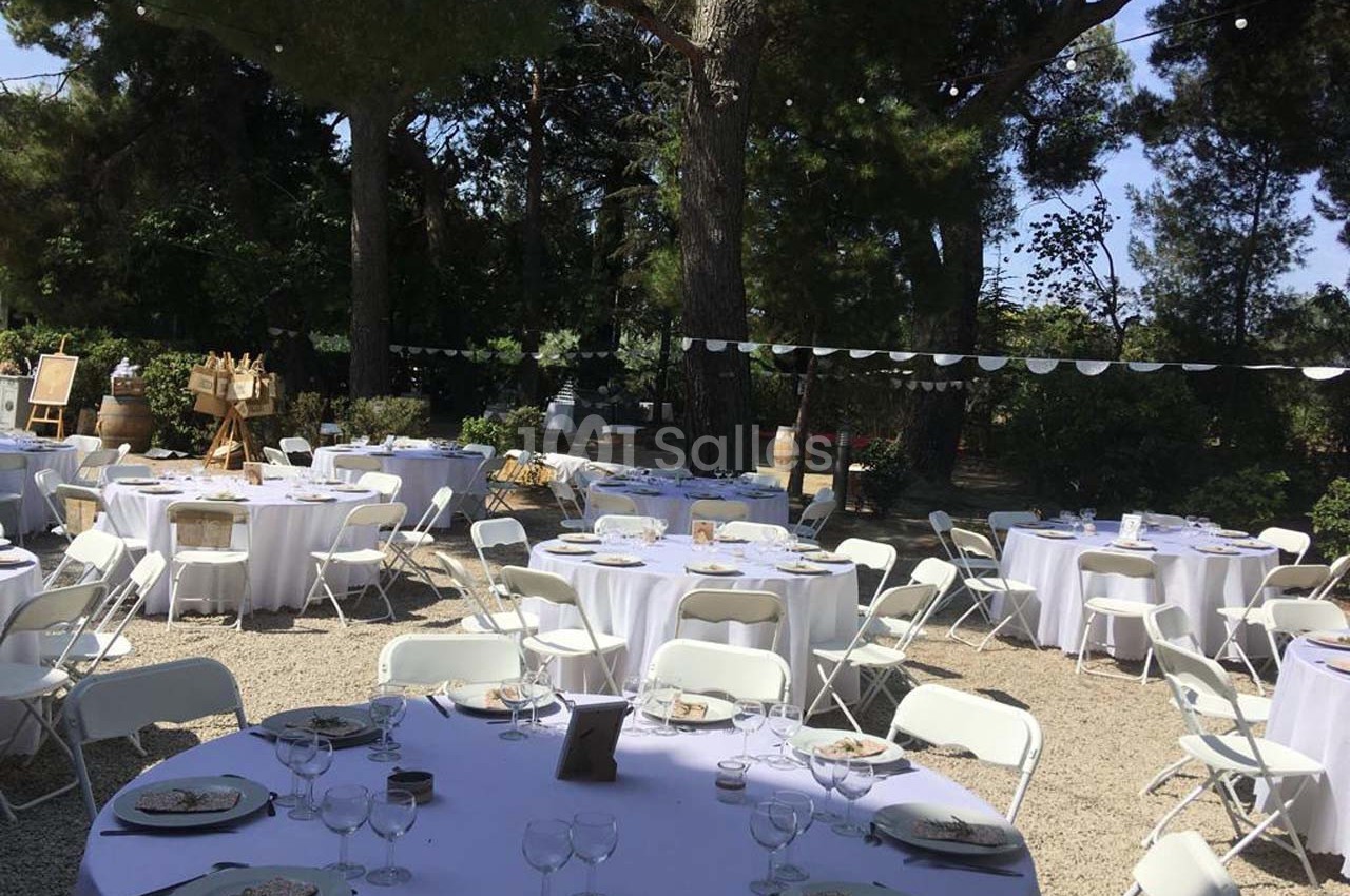 Tables rondes dressées avec nappes blanches et chaises pliantes dans un jardin ombragé, décoré pour un événement en plein…