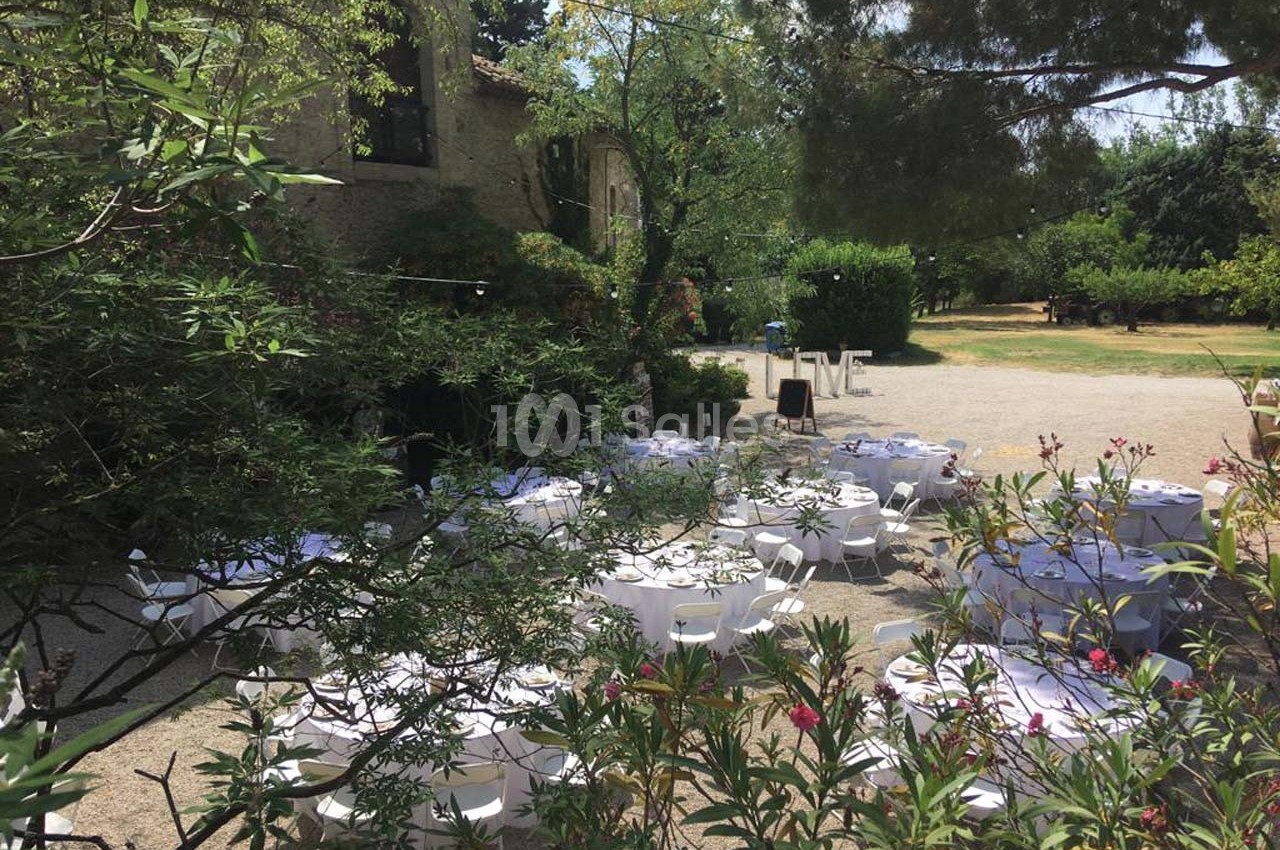 Tables rondes dressées en extérieur dans un jardin arboré, entourées de végétation, sous un ciel ensoleillé.