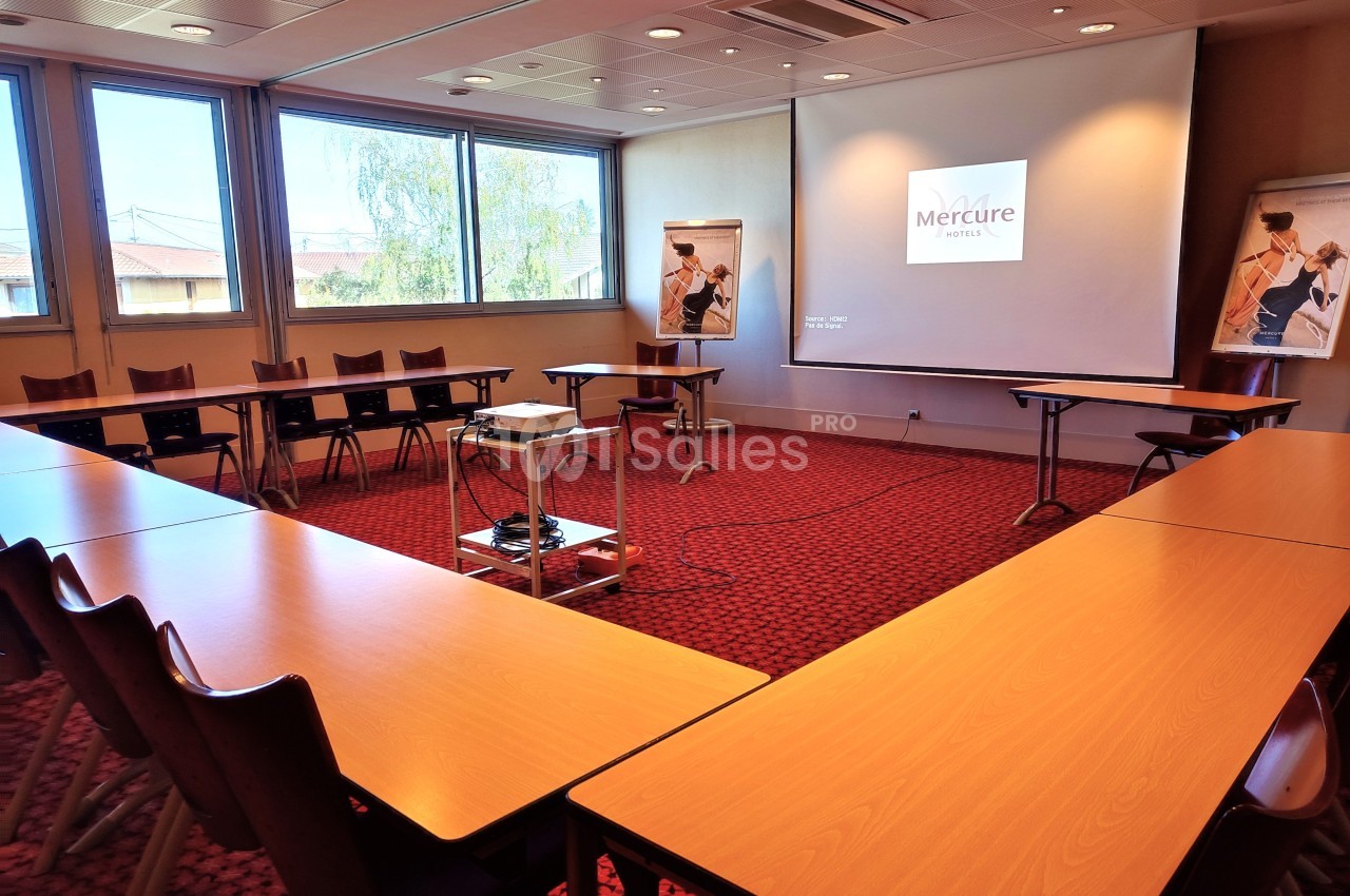 Salle de réunion lumineuse avec tables en U, projecteur allumé et affiches sur chevalets près des fenêtres.