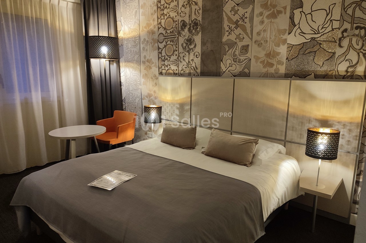 Chambre d'hôtel avec un lit double, deux lampes de chevet, un fauteuil orange et une table ronde près d'une fenêtre.