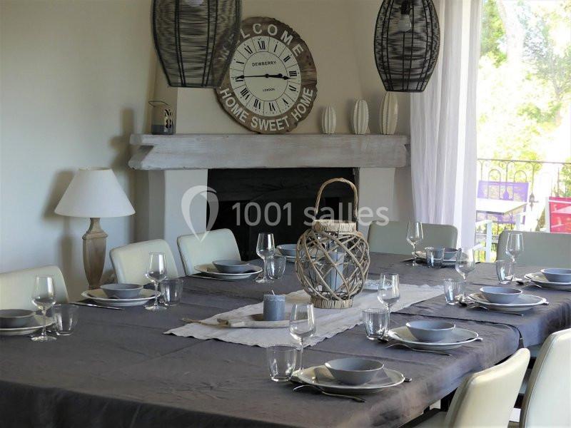 Table dressée avec vaisselle grise, nappe sombre et décoration en osier, dans une salle lumineuse avec cheminée.