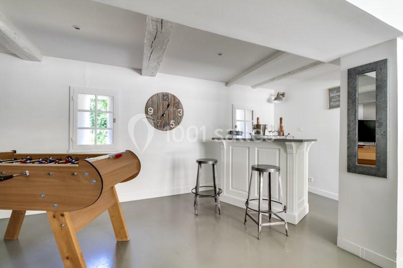 Salle lumineuse avec baby-foot en bois, bar blanc avec tabourets métalliques et horloge murale en bois.