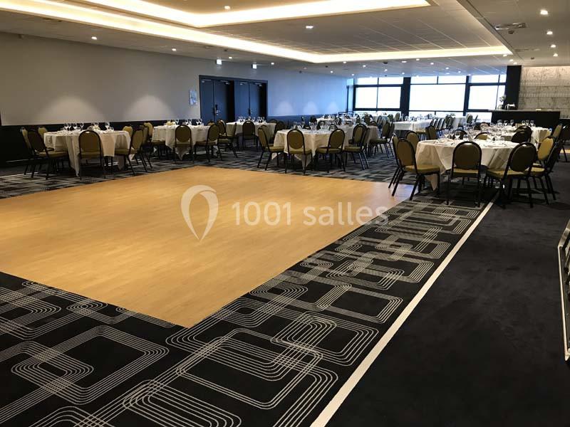 Salle de réception avec tables dressées autour d'une piste de danse en bois, éclairée par des lumières au plafond.