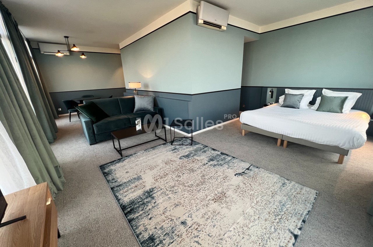 Chambre spacieuse avec lit double, canapé, tapis moderne, éclairage doux et murs aux tons verts et gris.