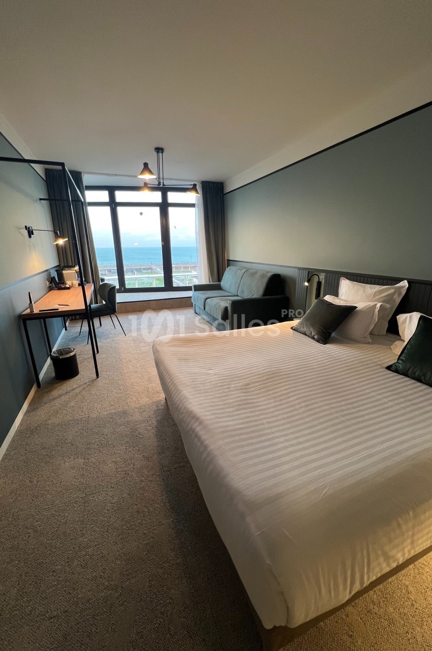 Chambre d'hôtel moderne avec lit double, bureau, canapé et grande fenêtre offrant une vue dégagée.