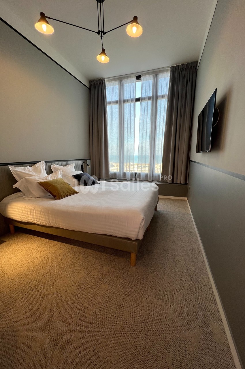 Chambre moderne avec lit double, grande fenêtre avec rideaux, éclairage suspendu et télévision murale.