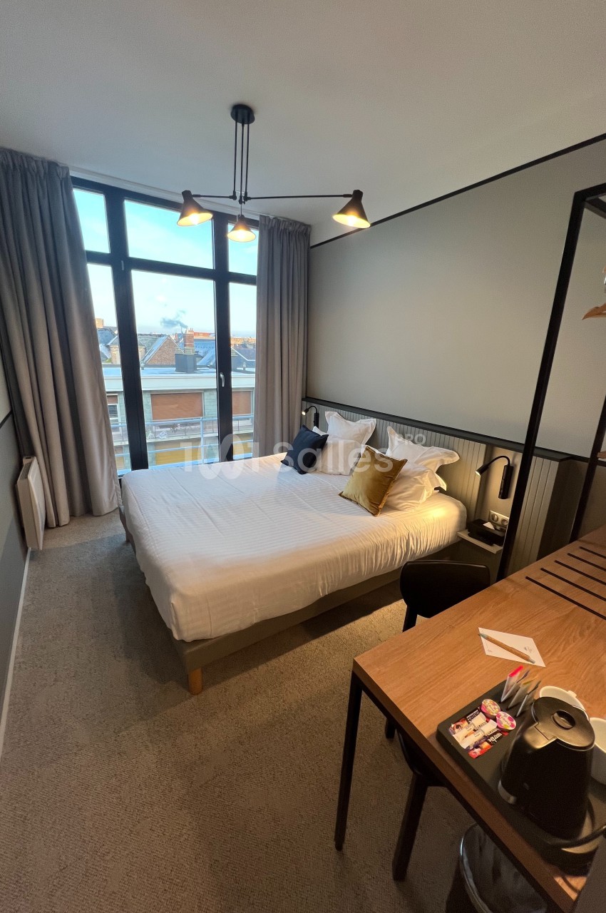 Chambre moderne avec lit double, bureau équipé, grande fenêtre avec vue sur les toits et éclairage suspendu.