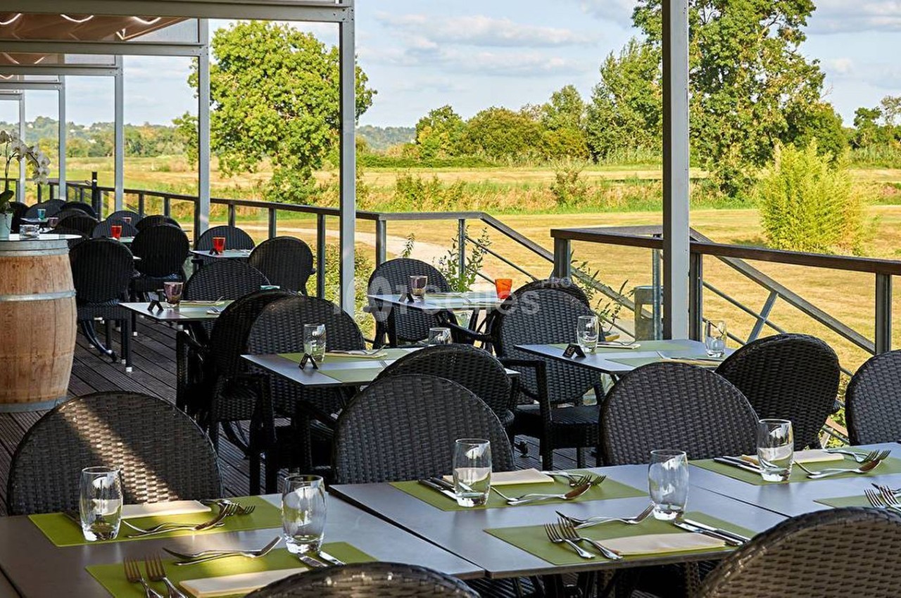 Terrasse d'un restaurant avec tables dressées, chaises en rotin noir et vue sur un paysage verdoyant.