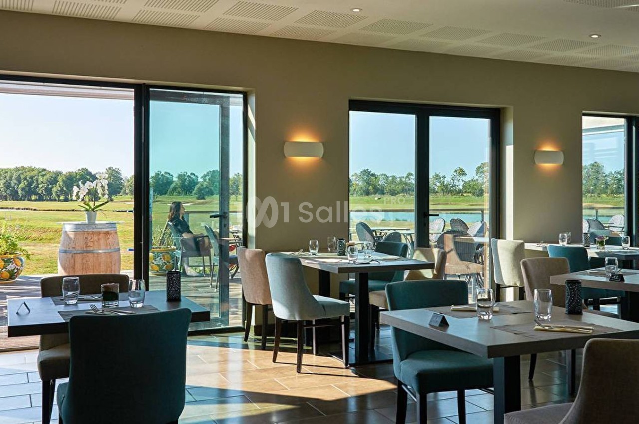 Salle de restaurant lumineuse avec grandes baies vitrées donnant sur une terrasse et un paysage verdoyant.