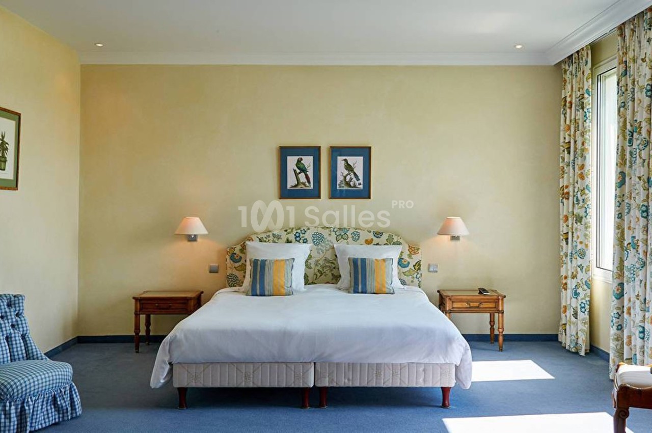 Chambre lumineuse avec lit double, deux tables de chevet, lampes murales, fauteuil et rideaux à motifs floraux.