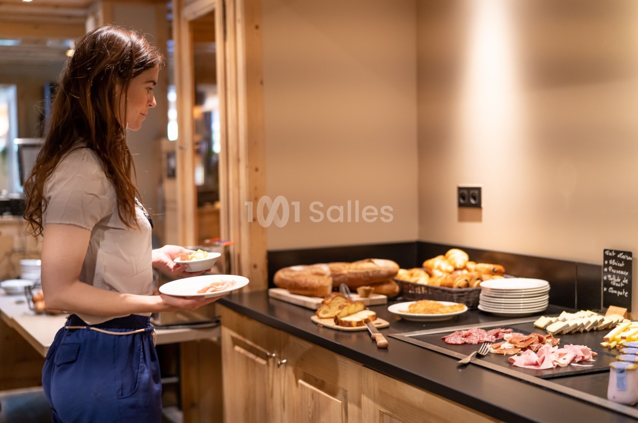Une femme se sert au buffet d'un petit-déjeuner avec pains, viennoiseries, charcuterie et fromages dans un cadre chaleureux.