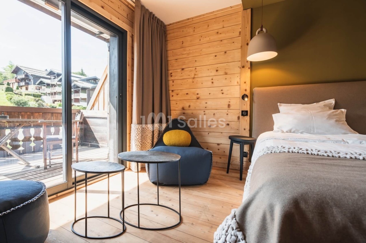 Chambre en bois avec lit, fauteuil, tables basses et balcon donnant sur des chalets en montagne.