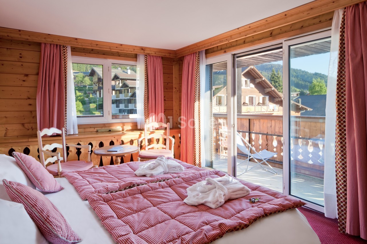 Chambre en bois avec lit double, balcon, grandes fenêtres et vue sur des chalets en montagne.