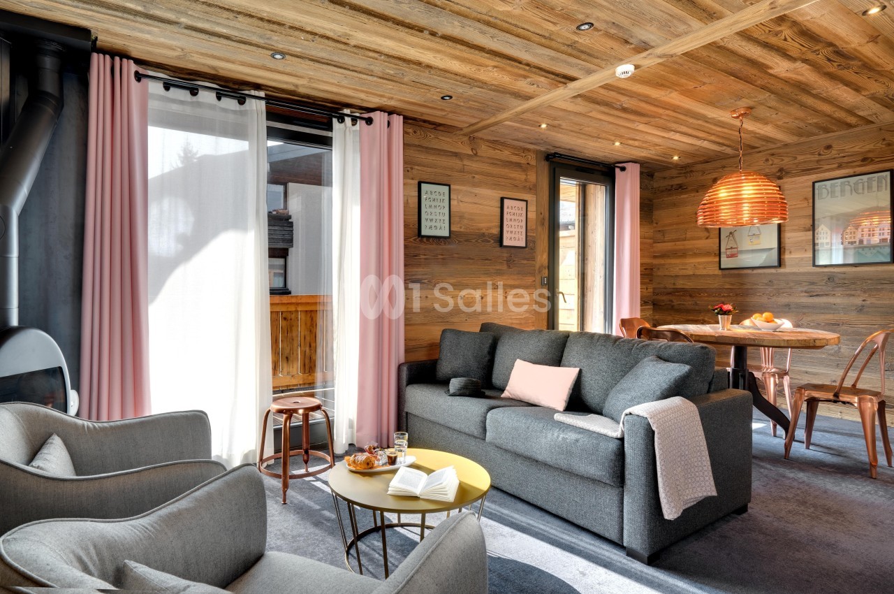 Salon chaleureux avec canapé gris, fauteuils, table basse, décoration en bois et lumière naturelle entrant par les fenêtres.