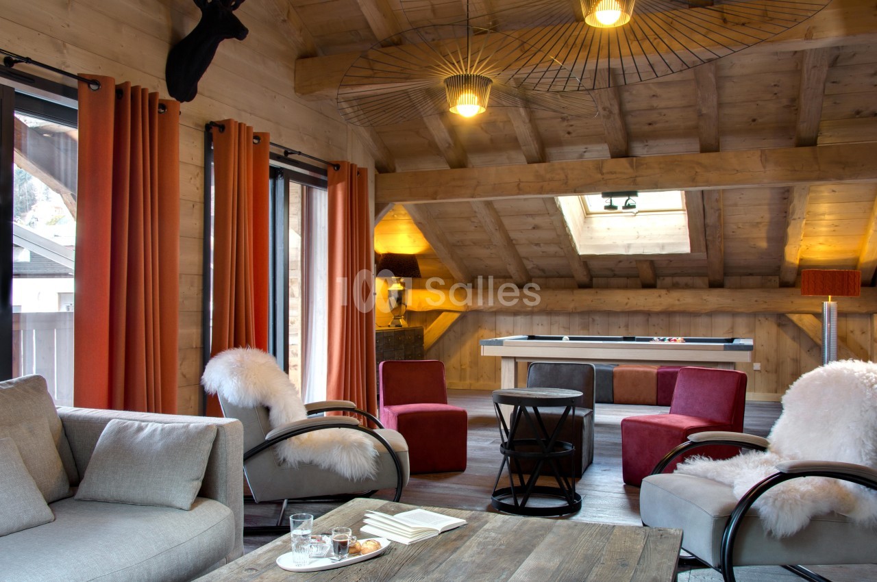 Salon chaleureux sous combles avec poutres apparentes, canapé, fauteuils, table basse en bois et table de billard.