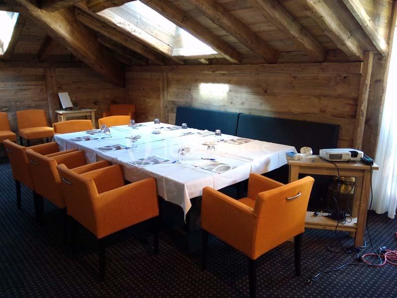 Salle de réunion avec table rectangulaire dressée, chaises orange, plafond en bois et projecteur sur un meuble.