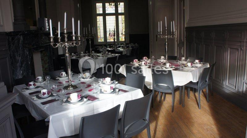 Salle de réception avec tables dressées, nappes blanches, chandeliers et vaisselle pour un repas ou événement.