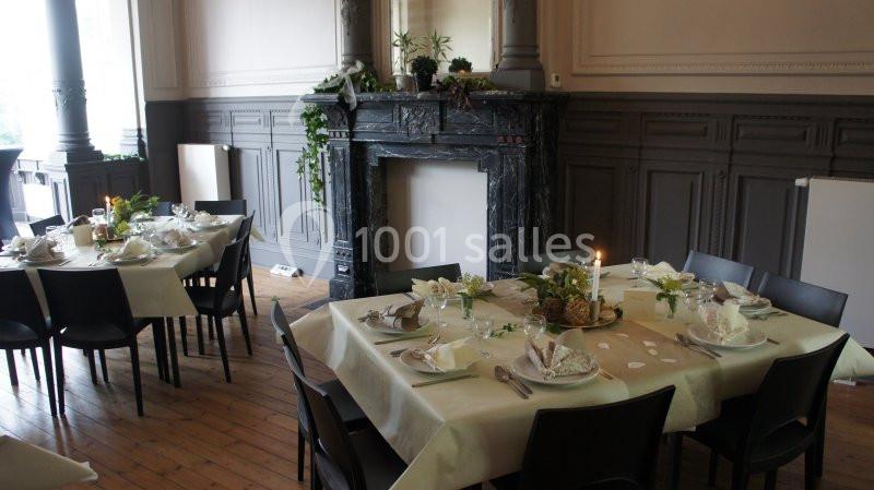 Salle élégante avec tables dressées, nappes claires, décorations florales et cheminée en marbre noir en arrière-plan.